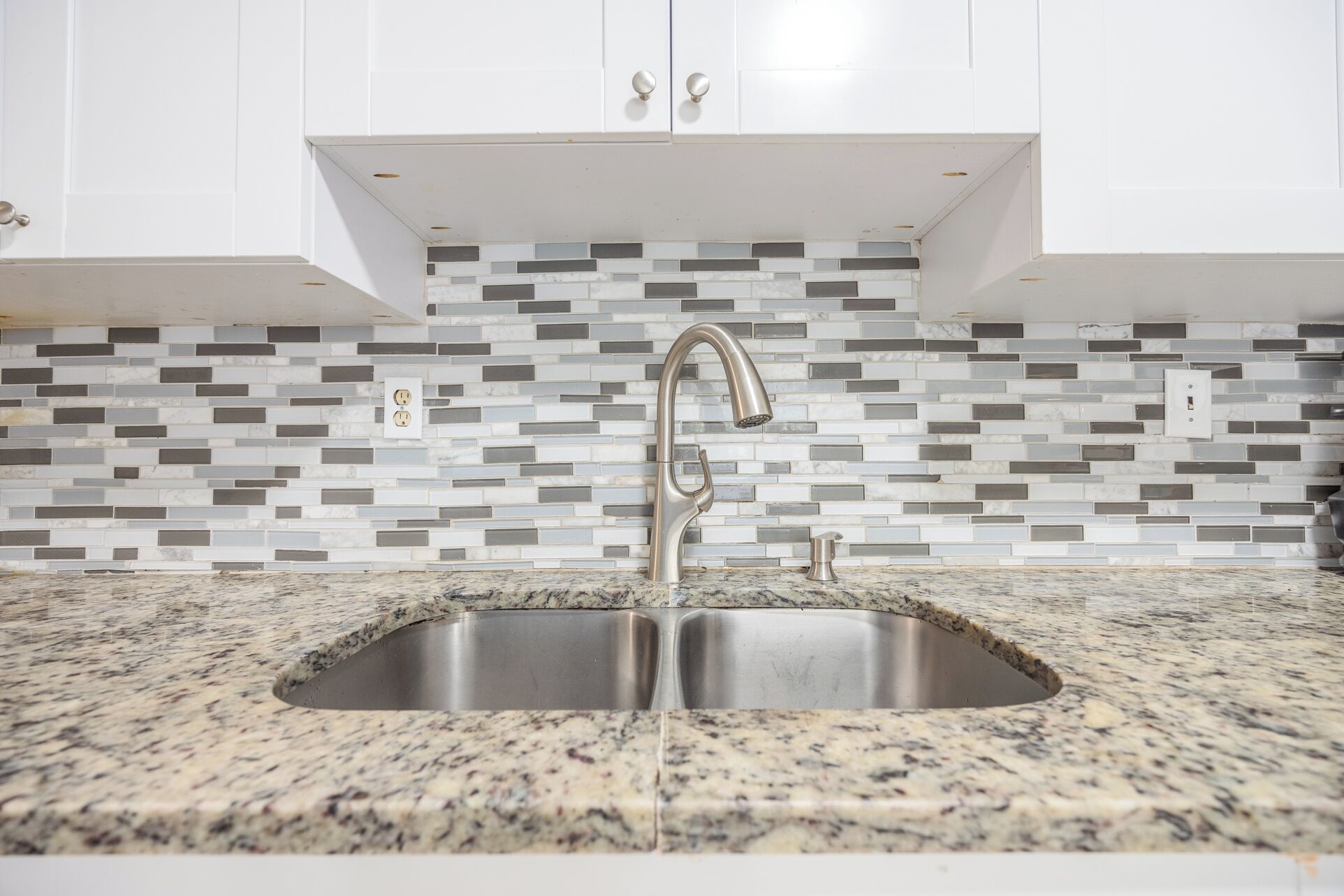 A kitchen with a double sink, granite countertop, white cabinets, and a mosaic backsplash.