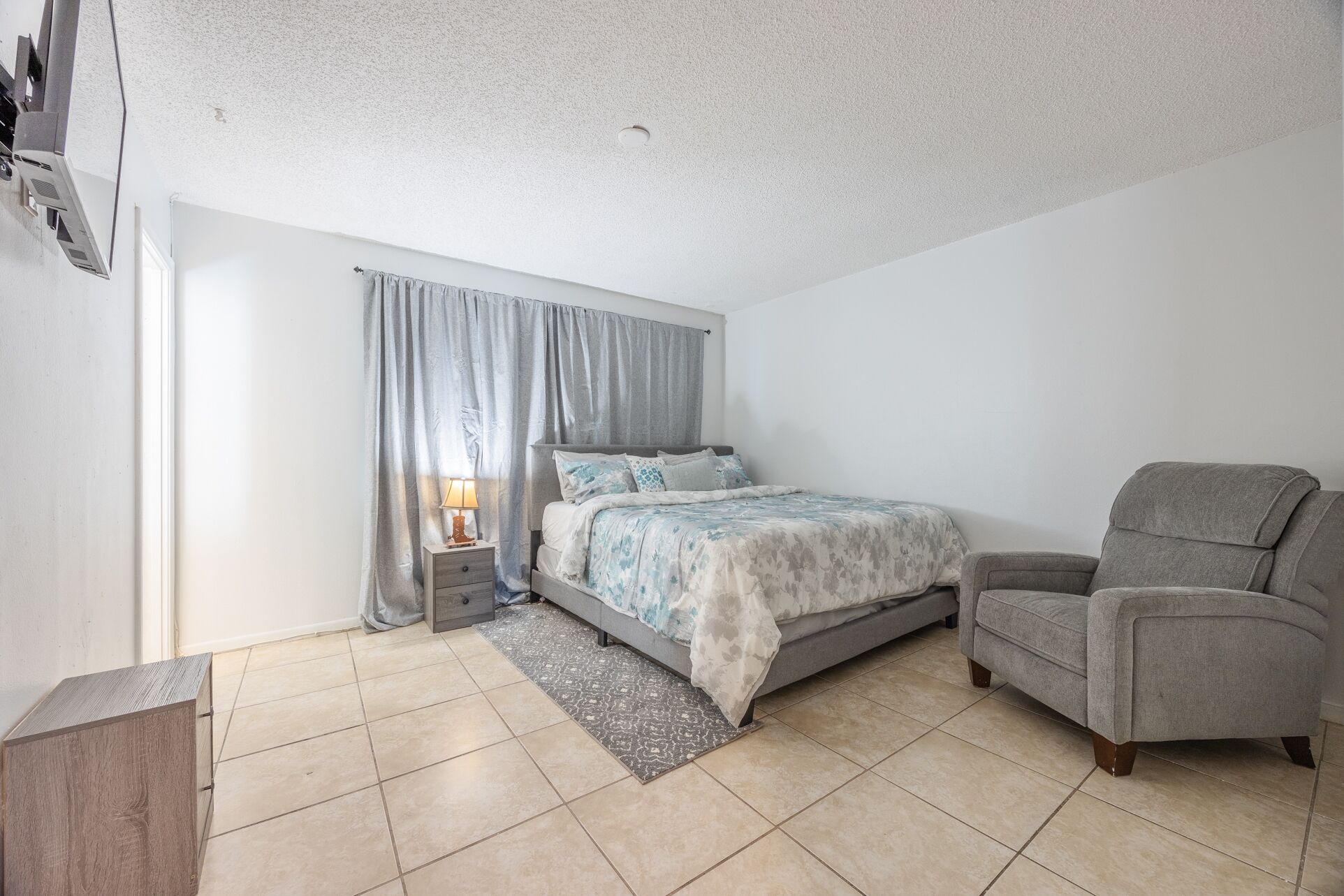 A bedroom with a bed, a chair, a nightstand with a lamp, and a window with curtains.