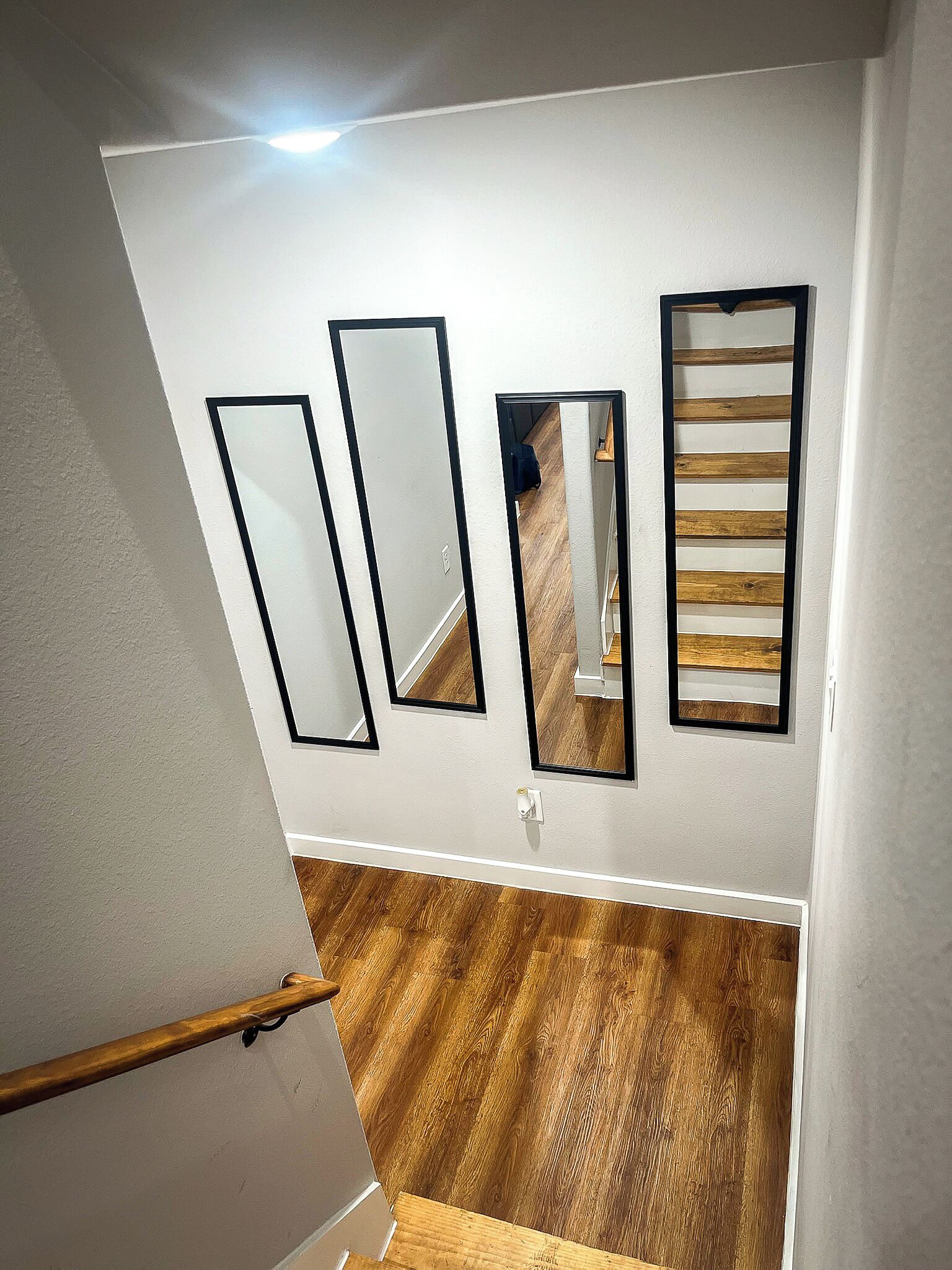 A staircase with a wooden floor and white walls. There are three mirrors on the wall, one of which reflects a wooden floor.