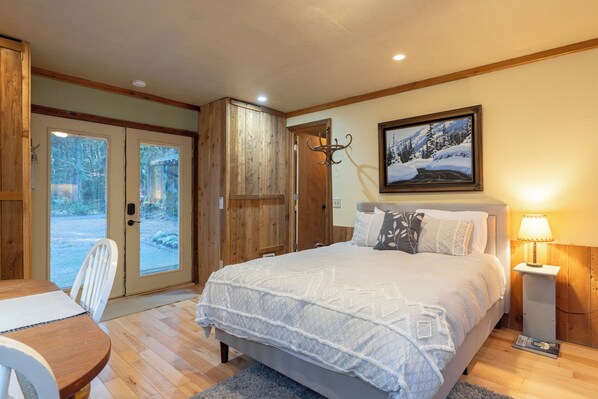 A bedroom with a large bed, a wooden wardrobe, a dining table, and a door leading to a balcony.