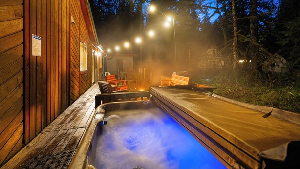 Een houten chalet met een hottub, sfeerverlichting en uitzicht op bomen in de schemering.
