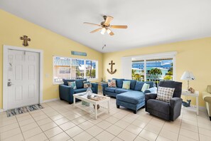 A living room with a blue sofa set, a ceiling fan, and a view of a blue building through the windows.