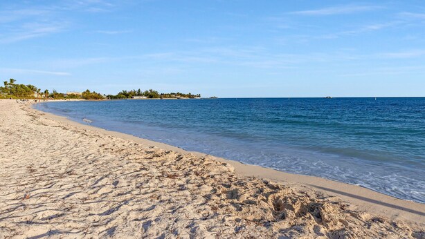 A beach with clear blue water, sandy shore, and a distant shoreline with buildings and trees. - Marathon 2BR | Private Pool & 24' Dock (Marathon)