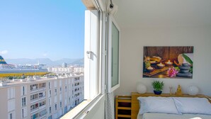 A balcony with a city view, a bed with white bedding, a painting on the wall, and a small Buddha statue. - Apartment L'Onyx by Interhome (Toulon)