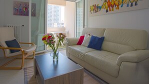 A living room with a beige sofa, a wooden coffee table with a vase of flowers, a white armchair, and a glass door leading to a balcony. - Apartment L'Onyx by Interhome (Toulon)