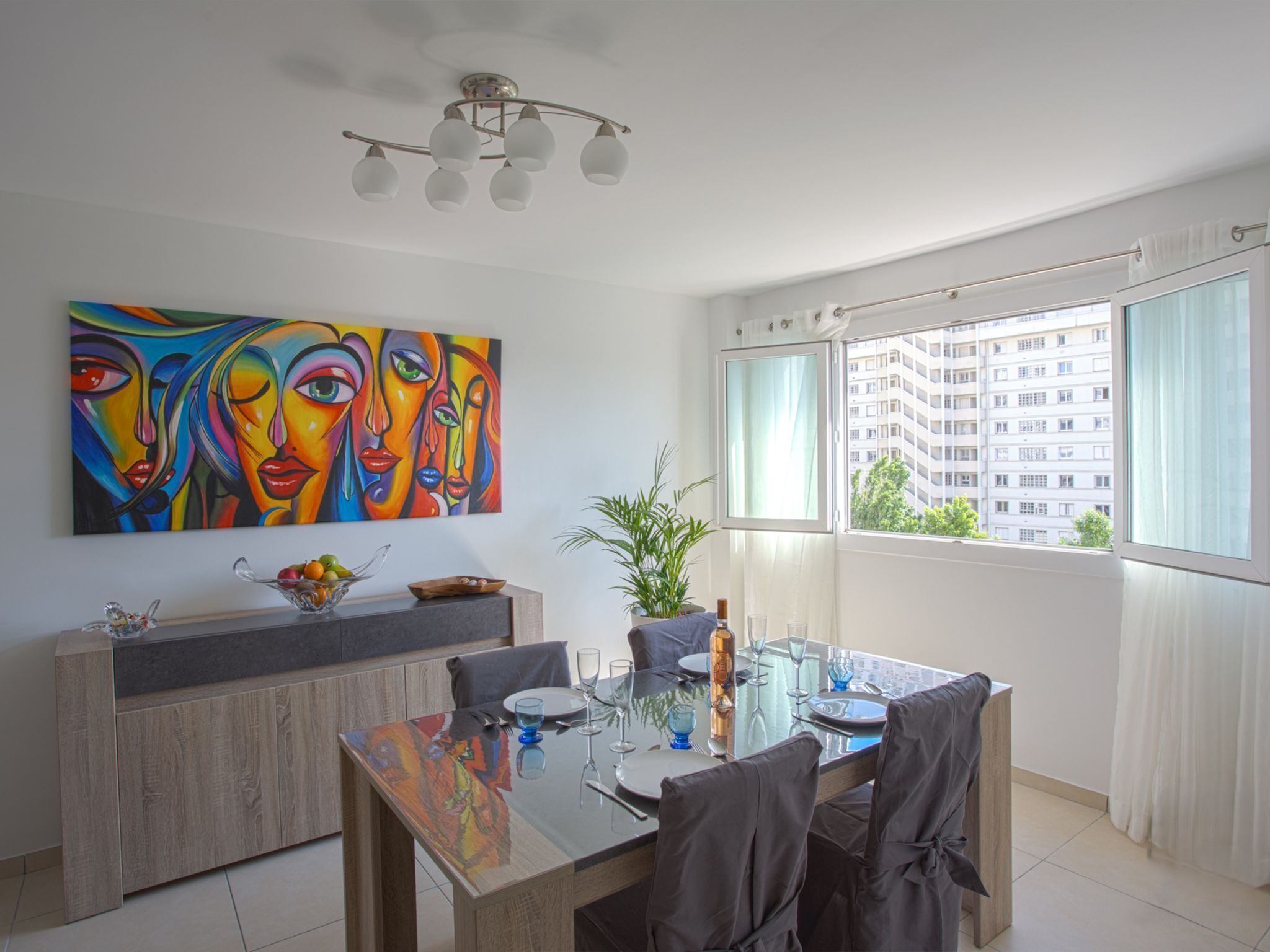 Zona pranzo con tavolo moderno apparecchiato per quattro persone, un quadro astratto dai colori vivaci sulla parete e vista su un edificio attraverso una finestra.