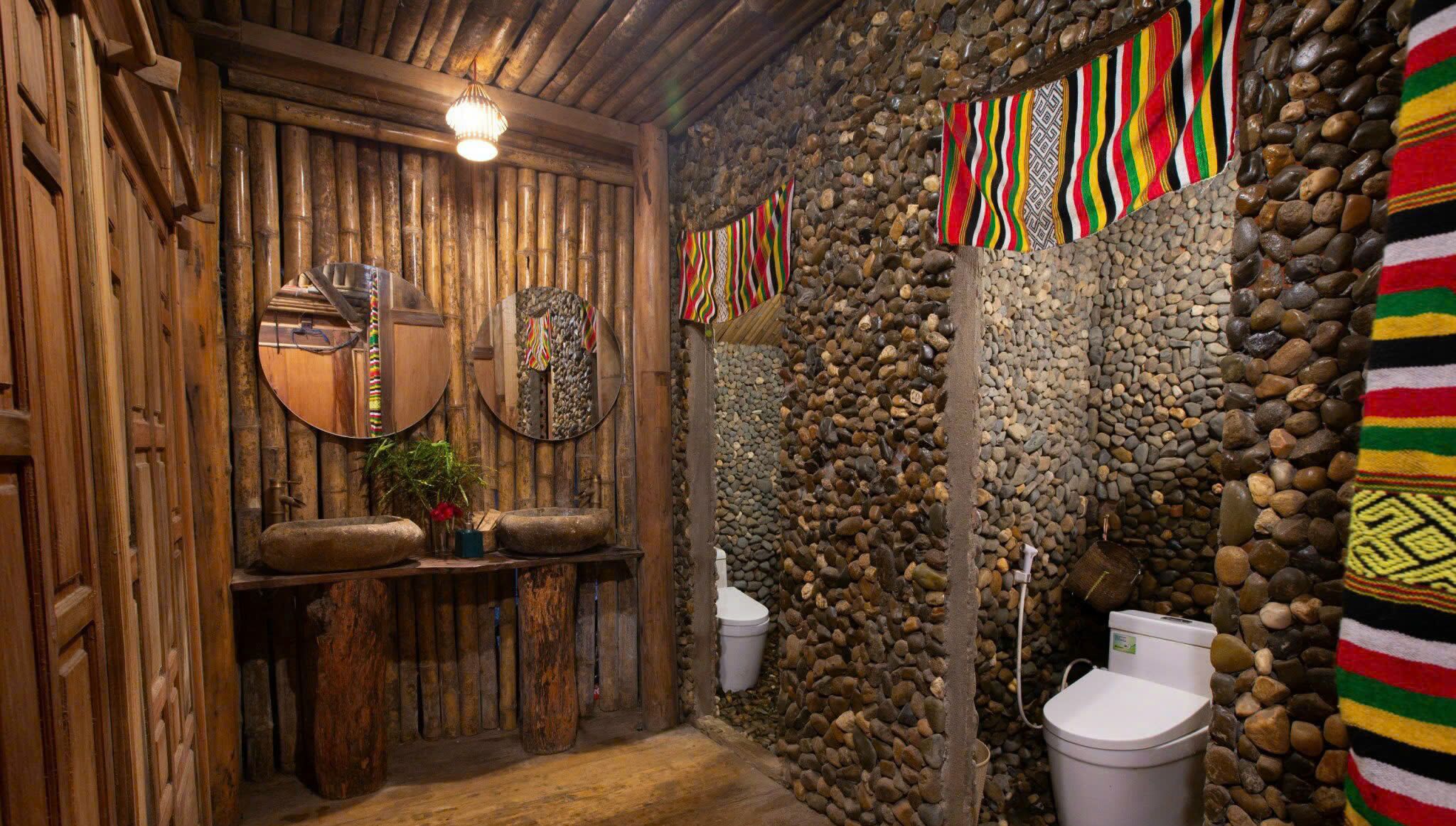 A rustic bathroom with wooden walls, a stone wall, and a wooden door. There are two toilets, a sink, and a mirror.