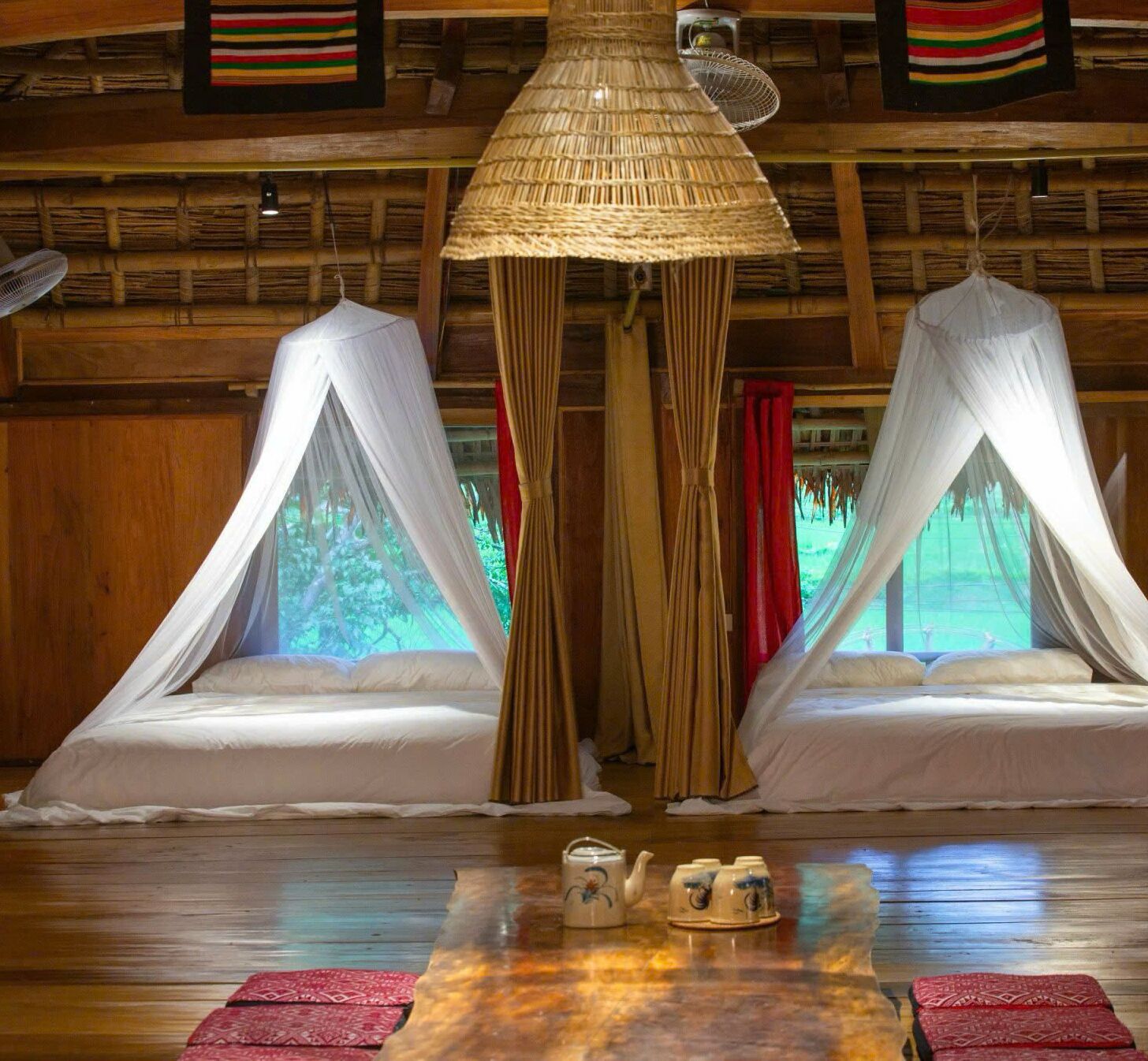 Una habitación con dos camas cubiertas por dos doseles blancos, una lámpara de techo de fibra natural, paredes de madera y vistas a la vegetación exterior.