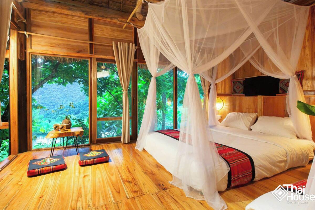 A room with a wooden floor, a bed with white bedding, a canopy bed, and a view of greenery outside.