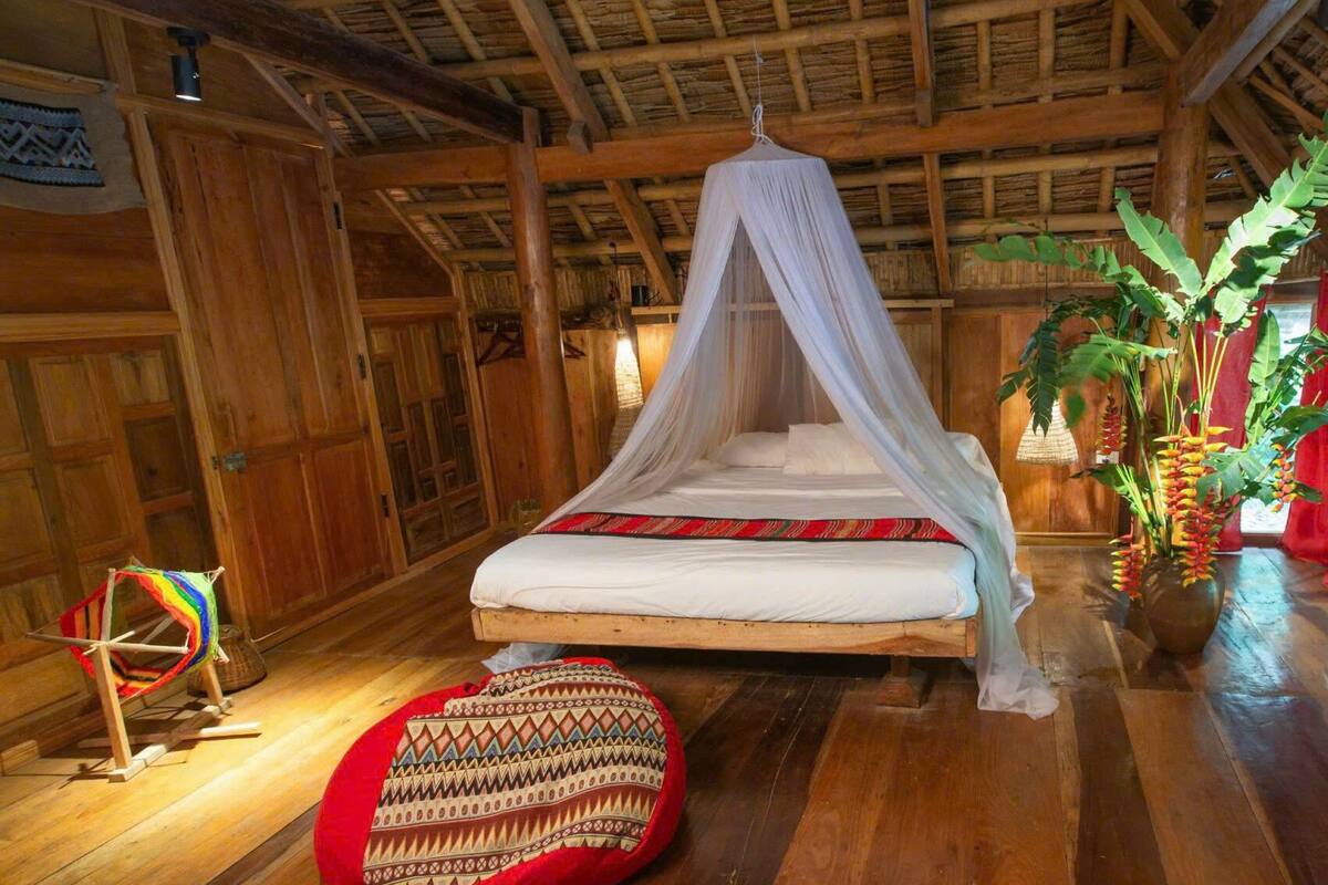 A traditional wooden room with a bed under a canopy, a red woven pouf, and a hanging plant.