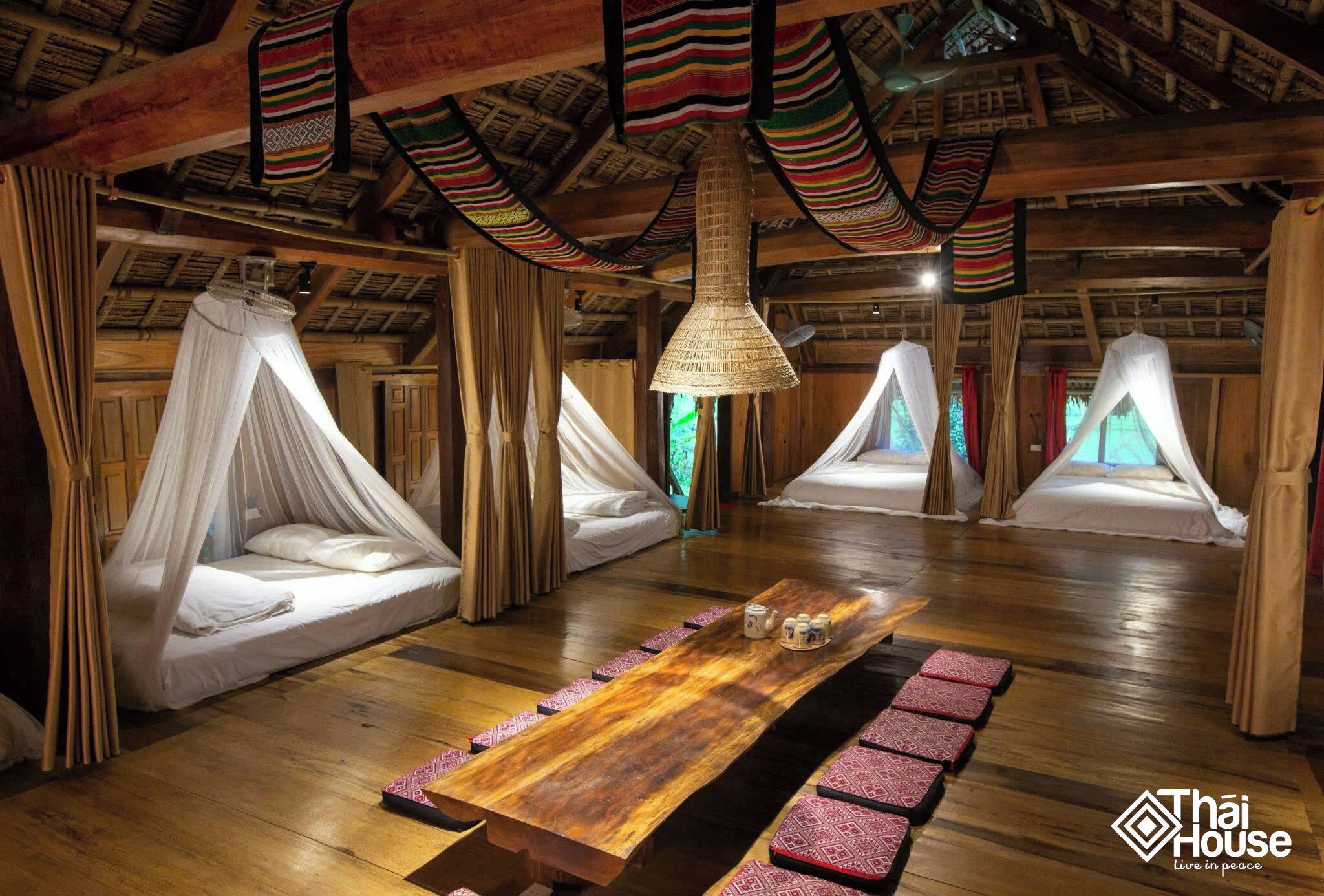 A room with wooden floors, a central wooden table, and four beds with canopy beds.
