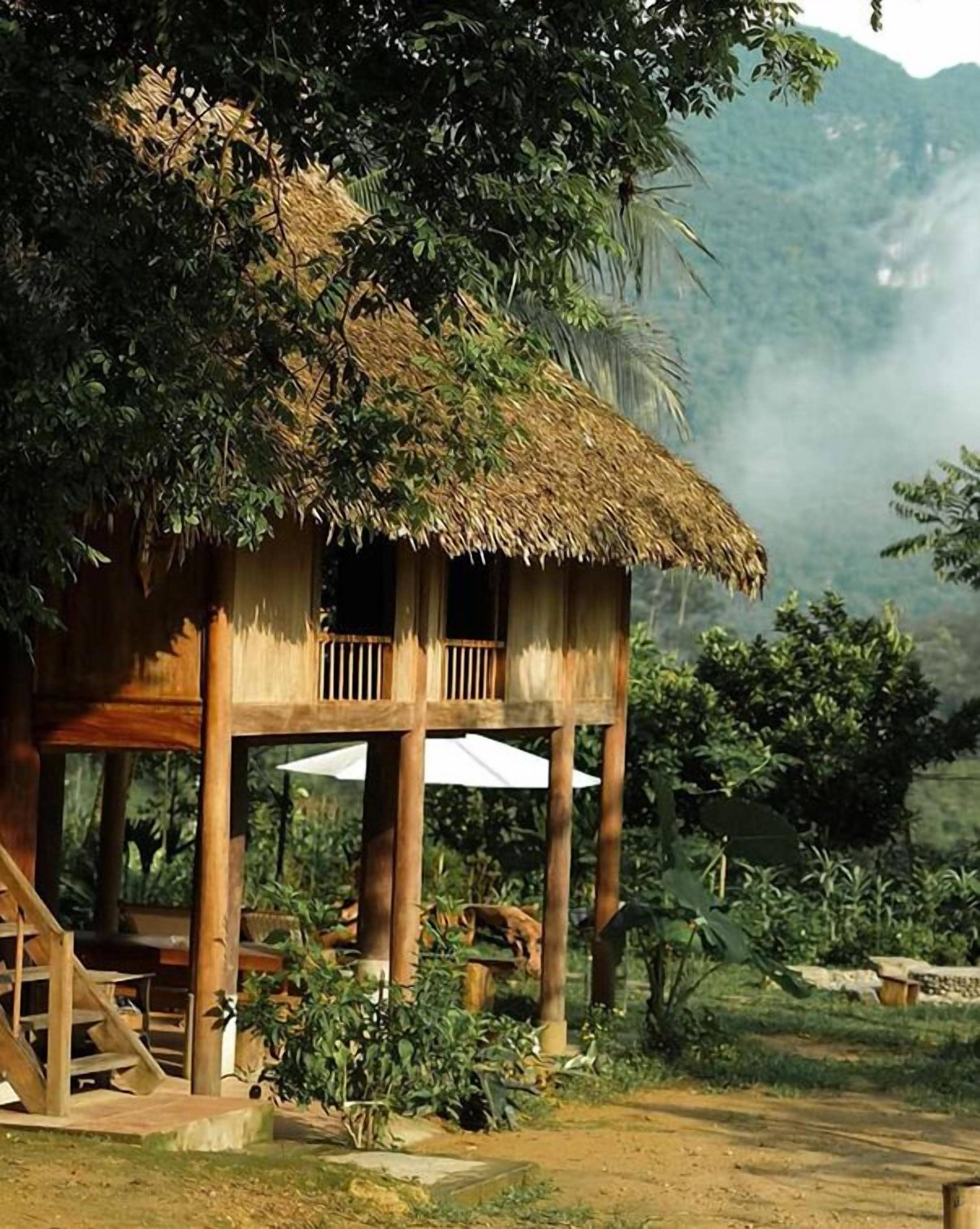 A thatched-roof wooden structure with a balcony, surrounded by lush greenery and a mountainous backdrop.