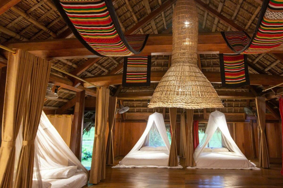 A room with a thatched roof, wooden beams, and a central woven light fixture. There are two beds with white linens and a view of greenery outside.