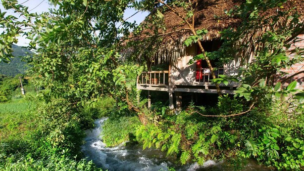 A rustic wooden bridge over a flowing river, surrounded by lush greenery and a thatched-roof structure. - Thai House Pu Luong (Co Lung)