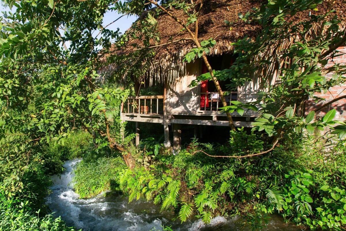A rustic wooden bridge over a flowing river, surrounded by lush greenery and a thatched-roof structure.