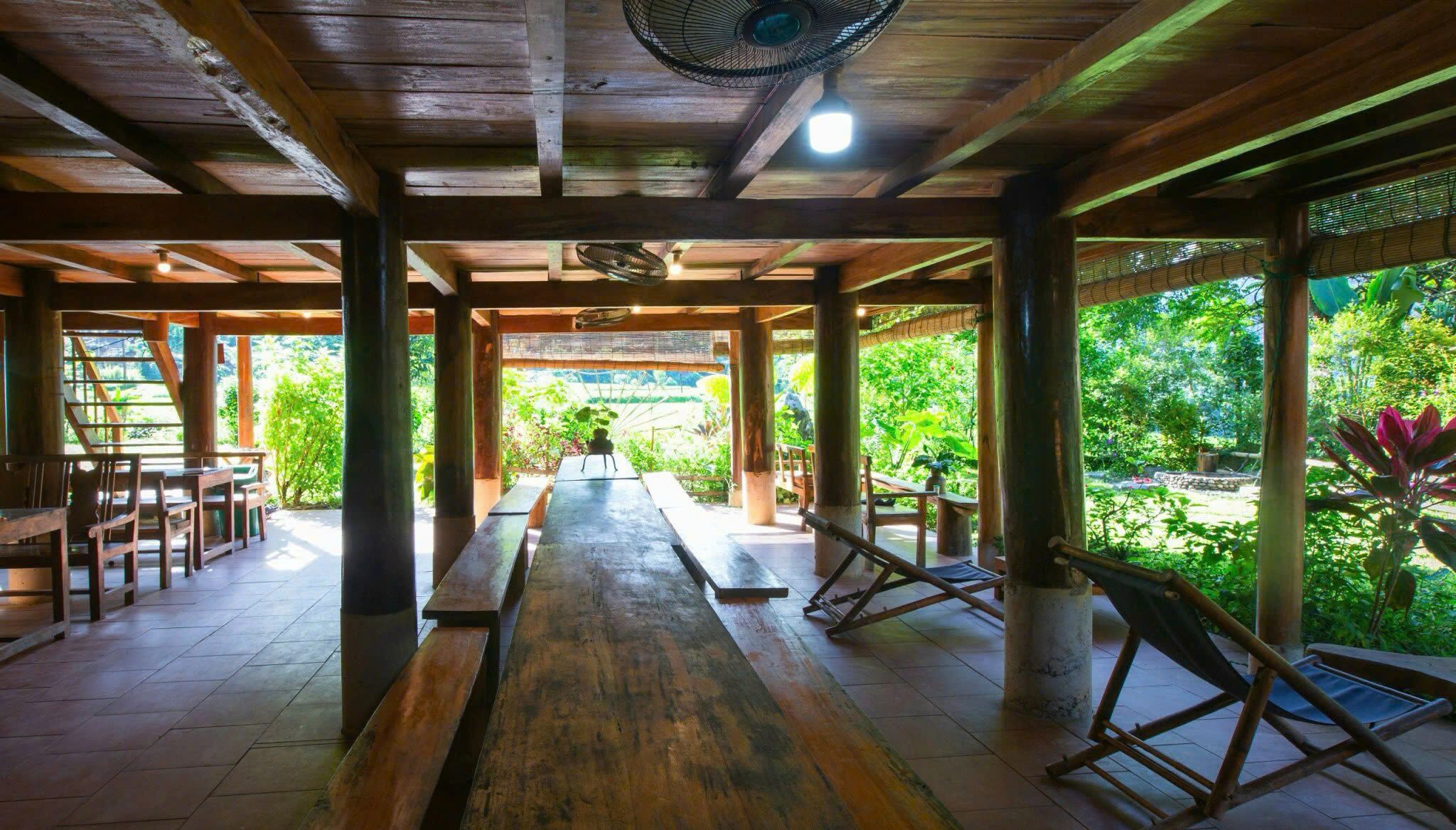 A covered outdoor area with wooden benches and tables, a fan, and a view of a lush garden.