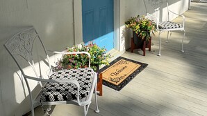 Varanda com porta azul, tapete de boas-vindas, duas cadeiras brancas e plantas em vasos.