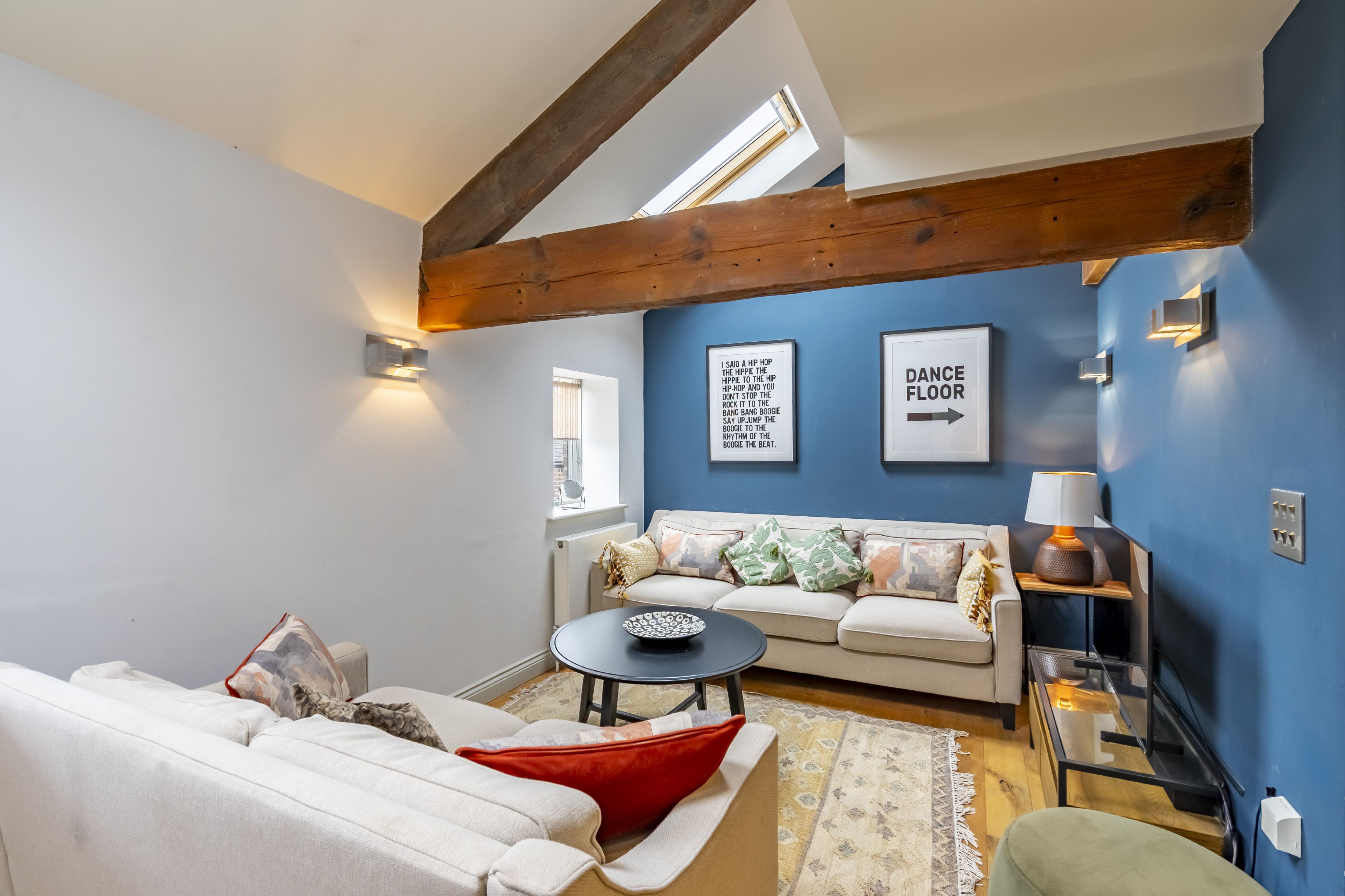 Ein Wohnzimmer mit einer Couch, einem Couchtisch und einer Lampe. An der Wand hängen zwei gerahmte Bilder und es gibt ein Fenster mit Jalousien.