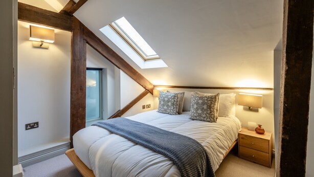 Une chambre avec un puits de lumière, un lit agrémenté d’oreillers à motifs, une table de chevet avec une lampe et une tête de lit en bois.