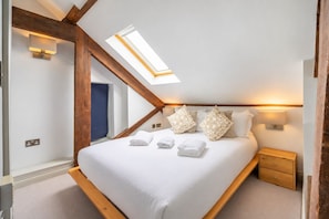 Ein Schlafzimmer mit schrägem Dach, einem Oberlicht, einem Bett mit weißer Bettwäsche, gemusterten Kissen, Nachttischlampen und einem Holznachttisch.