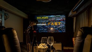 Un système de cinéma maison avec un grand écran affichant les titres « BELFAST » et « FAST », une bouteille de vin, du pop-corn et deux verres posés sur une table.