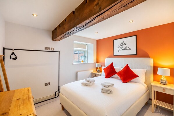 Habitación de hotel con una cama grande, una viga de techo de madera, una ventana con persianas, un cartel enmarcado en la pared y una mesita con una lámpara.