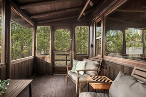 A wooden deck with a sofa, a small table with a chessboard, and large windows offering a view of trees. - Cozy w/Fire Pit, Grill, Screened Porch & WiFi (Harpers Ferry)