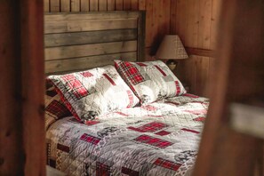 Ein hölzernes Bett mit gesteppter Bettdecke in Rot-Weiß-Mustern, ein Holz-Kopfschild und eine Nachttischlampe.