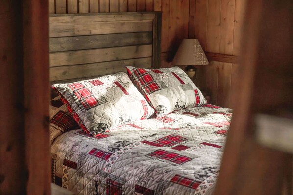 A wooden bed with a quilted bedspread featuring red and white patterns, a wooden headboard, and a bedside lamp.