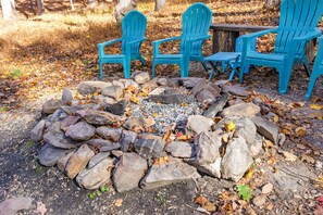 A fire pit surrounded by rocks and blue Adirondack chairs in a wooded area.