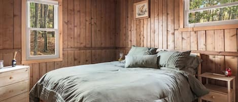 A wooden cabin bedroom with a bed, two bedside tables, a dresser, and two windows.
