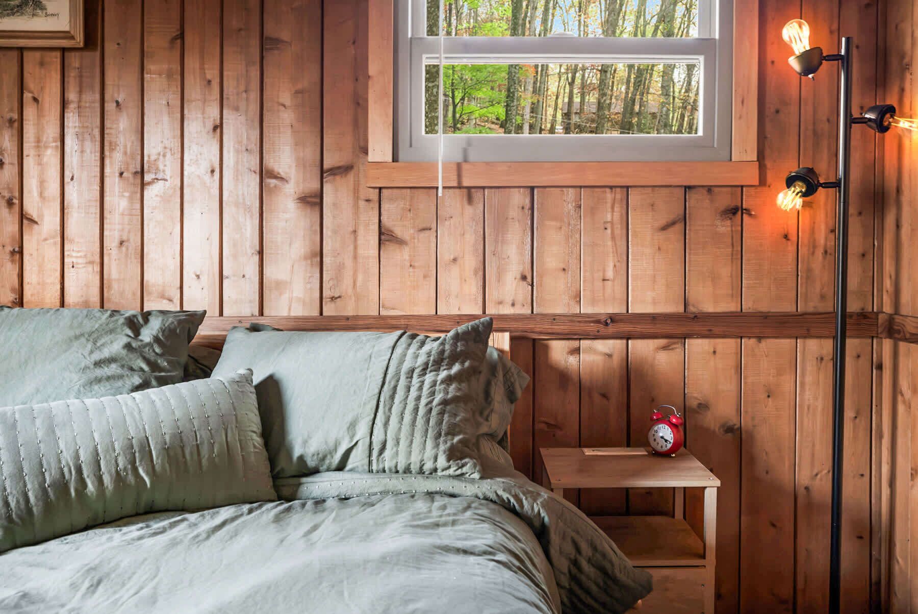 Une chambre rustique en bois, avec un lit, des oreillers, une table de chevet sur laquelle se trouve un réveil rouge, et une fenêtre donnant sur la forêt.