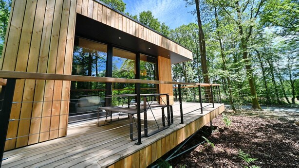 Una cabaña de madera moderna con terraza, ventanas amplias y vistas al bosque.