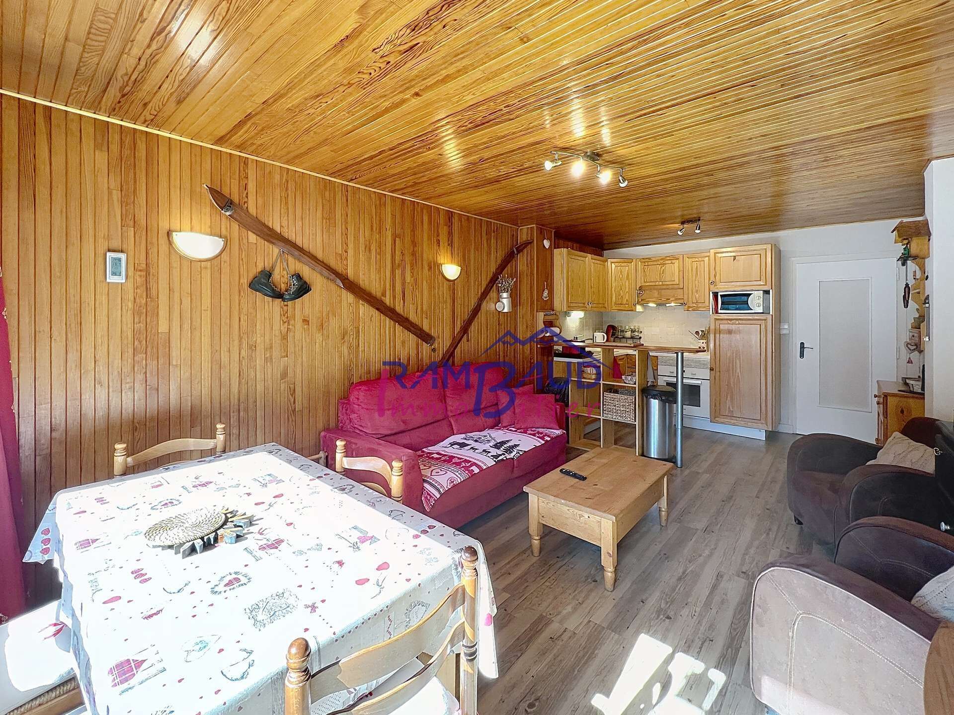 Ein gemütliches Blockhäuschen im Inneren mit Holzbalkendecke, einem Esstisch mit floralem Tischläufer, einem roten Sofa und einer Küchenzeile mit Holzausbau.