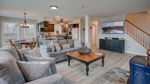 A spacious living room with a large gray sectional sofa, a wooden coffee table, and a stone accent rug. A dining area with a wooden table and chairs is visible in the background. The kitchen is modern with dark cabinets and stainless steel appliances. A large TV is mounted on the wall above the sofa. - Stunning Views | Hot Tub | Pets (Carroll)