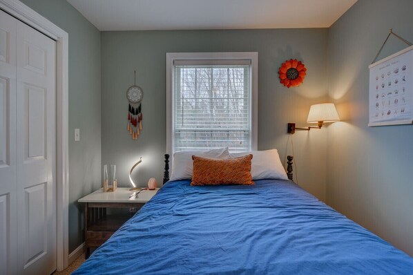 Une chambre à coucher avec un lit, une fenêtre, un attrape-rêves fixé au mur, une lampe murale et une affiche.