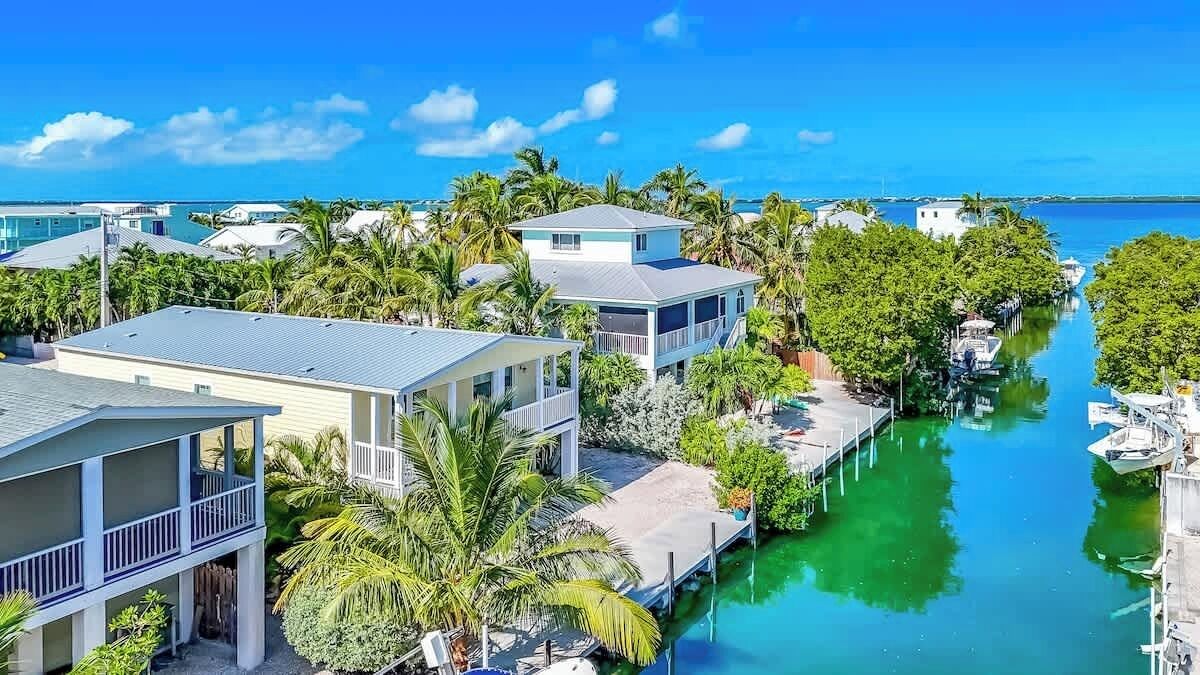 A coastal residential area with houses along a narrow waterway, surrounded by lush greenery and palm trees.