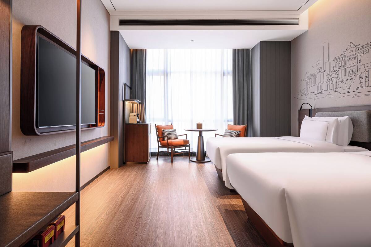 A hotel room with a large flat-screen TV, two beds, a small table with chairs, and a large window with curtains.