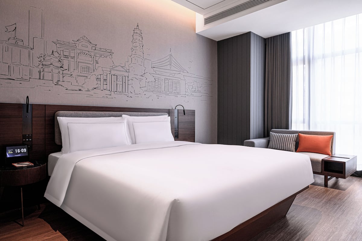 A modern hotel room with a large bed, a nightstand with a digital clock, a seating area with a chair and a small table, and a wall mural depicting traditional architecture.