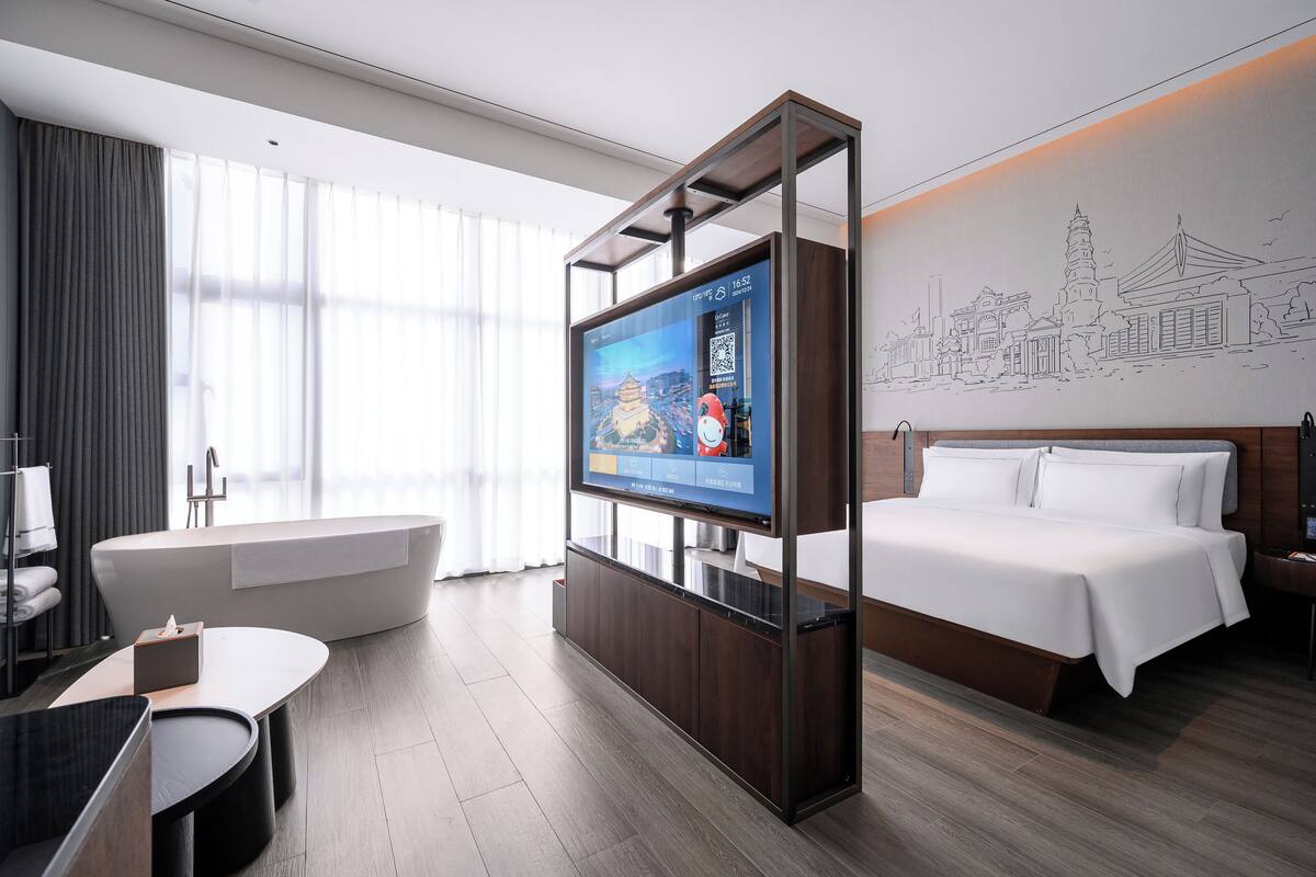 A modern hotel room with a large flat-screen TV, a bathtub, a bed, and a wall with a cityscape illustration.