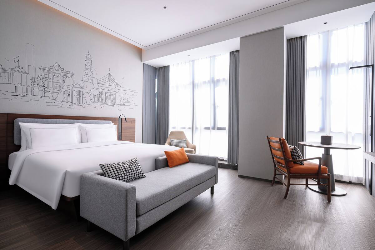A hotel room with a large bed, a grey sofa, a dining table with a chair, and a cityscape mural on the wall.
