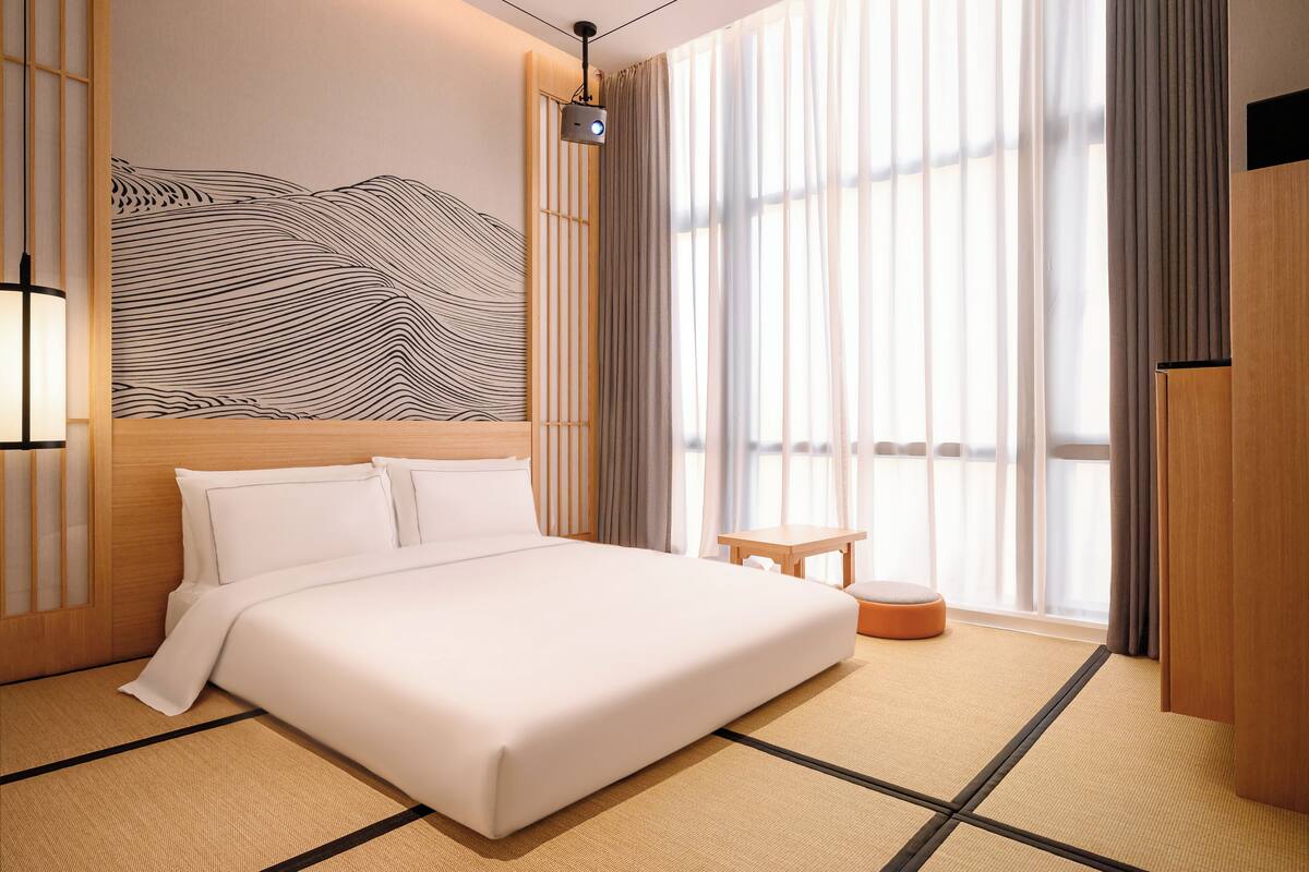 A hotel room with a large bed, a small table, a wall-mounted projector, and a view through large windows.