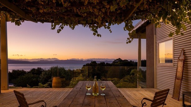 Eine Holzterrasse mit Tisch für Zwei, mit Blick auf eine malerische Meeres- und Berglandschaft bei Sonnenuntergang.