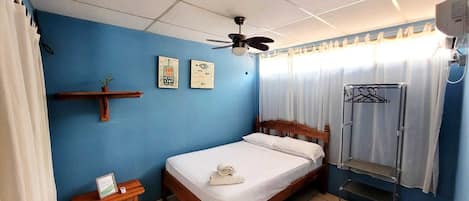 A bedroom with a wooden bed, a ceiling fan, a small table with a sign, and a wall-mounted shelf with a plant.