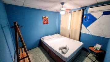 A small, single-bed room with a blue wall, a ceiling fan, a bed with a white mattress and pillows, a small table with a chair, and a painting on the wall.