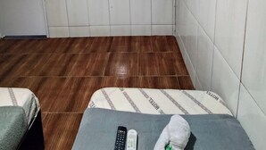 Una habitación de hotel pequeña con suelo de madera, una cama con colcha de cuadros grises y blancos, un control remoto, un peluche y un televisor montado en la pared.
