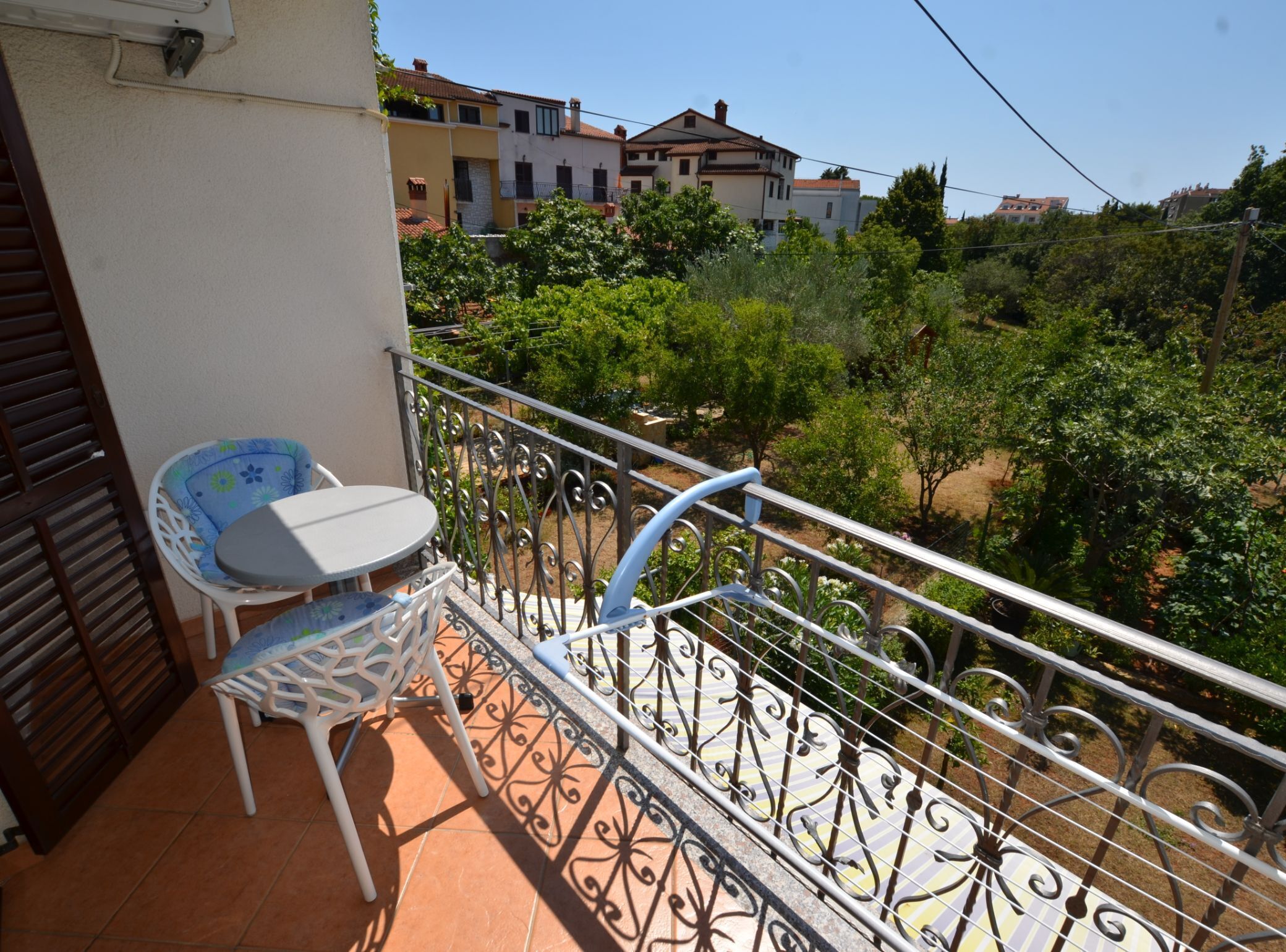 Ein Balkon mit Tisch und Stühlen, einem Metallgeländer und Blick auf umliegende Gebäude und Grünflächen.