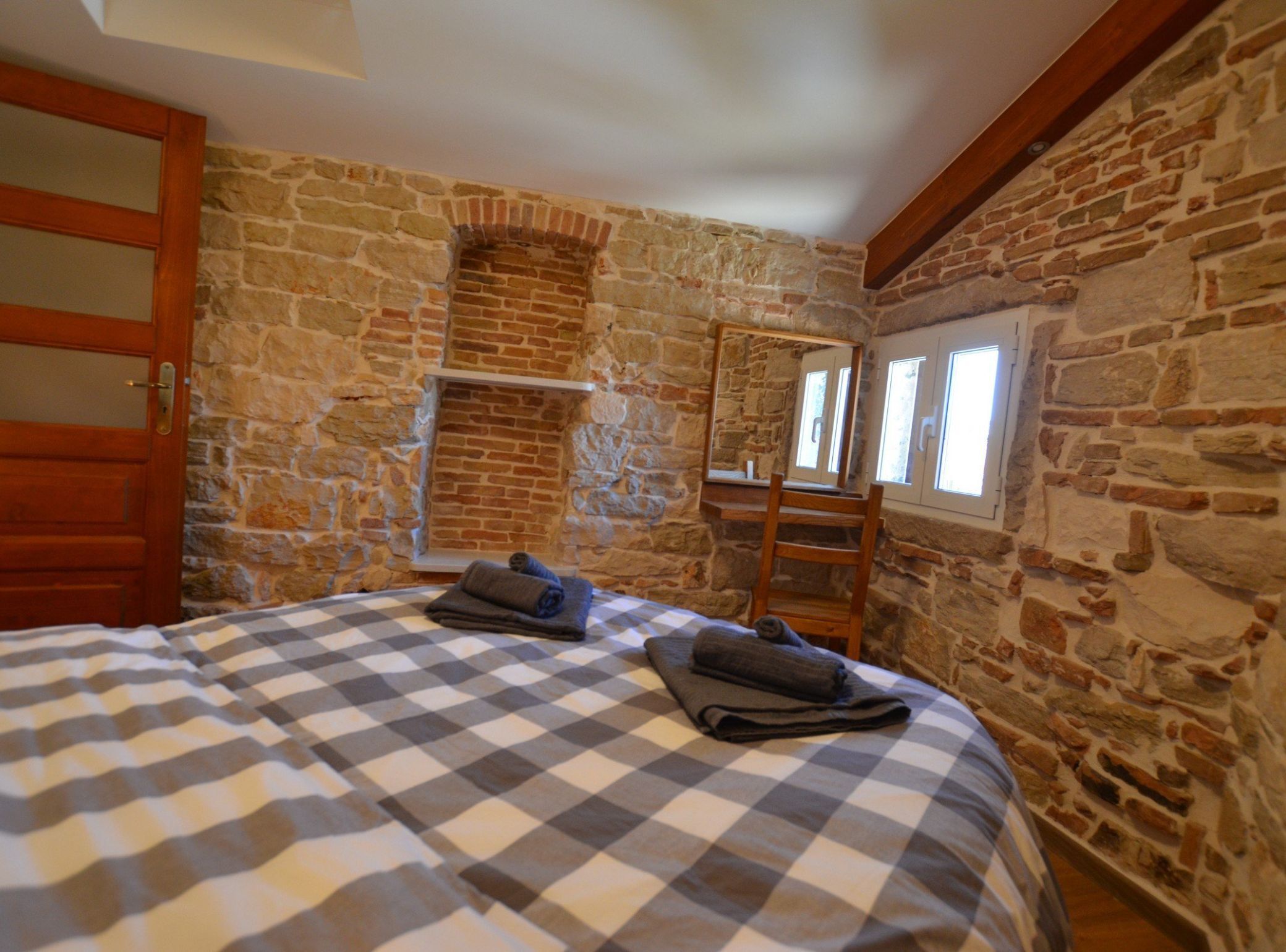 Een slaapkamer met een stenen muur, een houten deur, een geruit beddegoed en een spiegel aan de muur.