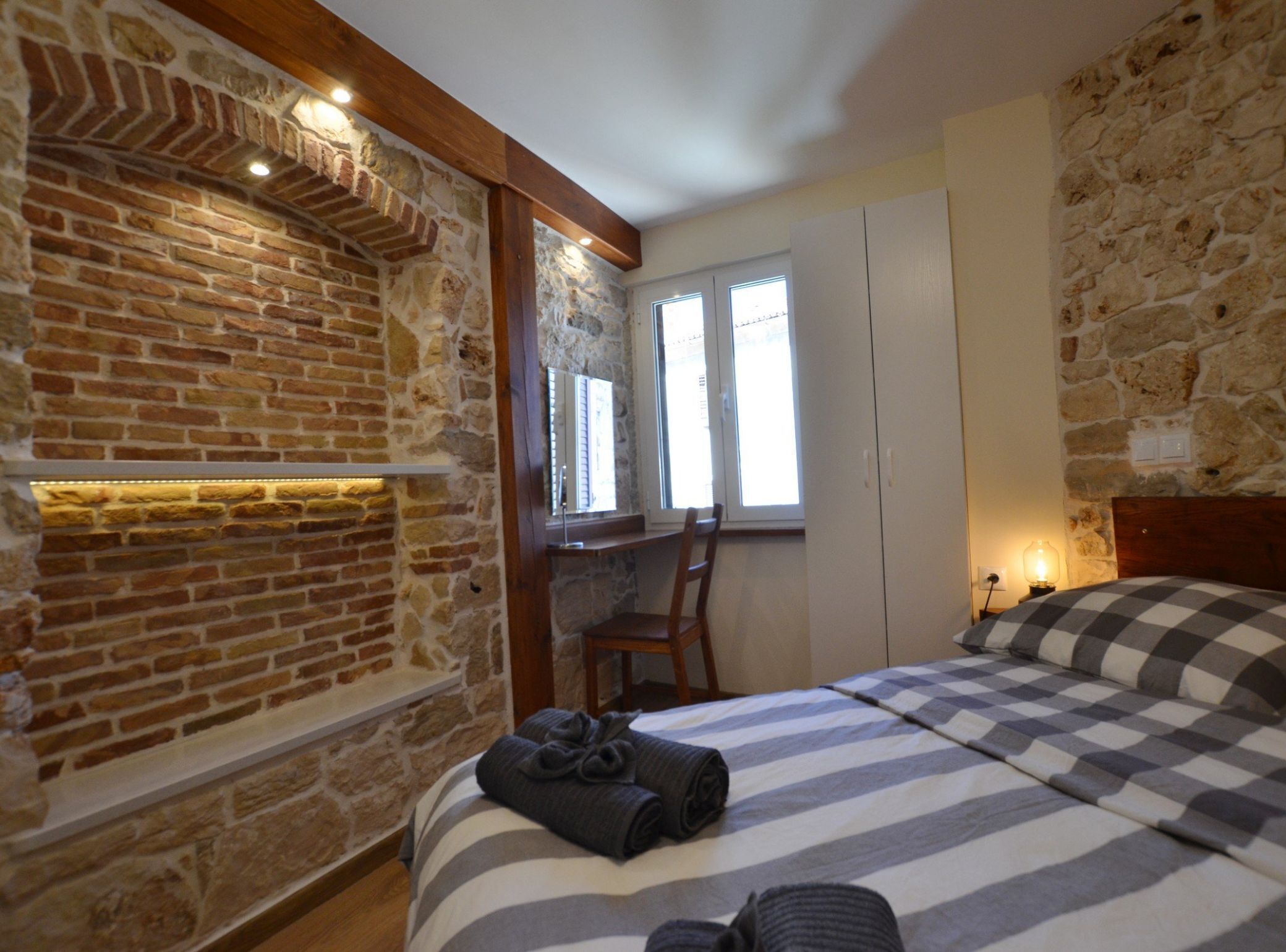 A bedroom with a stone wall, a bed with striped bedding, a wooden headboard, and a window with shutters.