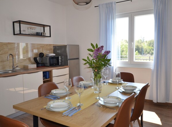 A dining area with a wooden table set for four, a kitchen in the background, and a window with curtains. - Lush and Sweet Escape Apartments (Rovinj)